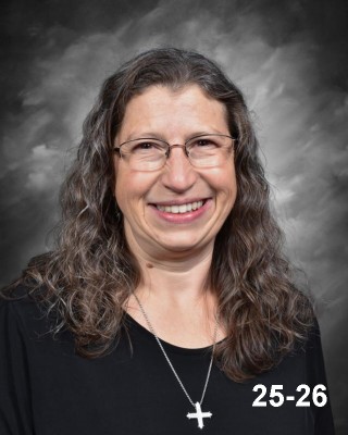 A smiling woman with long, curly hair and glasses, wearing a black top and a cross necklace, against a gray backdrop.