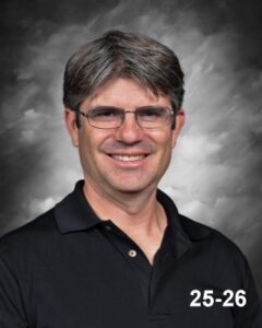 A man with short gray hair and glasses smiles, wearing a black polo shirt against a gray backdrop.