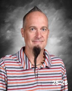 A man with a mohawk hairstyle and a long goatee smiles, wearing a striped shirt against a gray background.