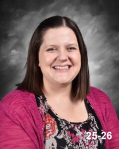 A woman with shoulder-length brown hair smiles, wearing a floral top and a pink cardigan, against a gray background.