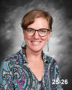 A smiling woman with glasses and earrings, wearing a colorful patterned shirt, against a gray background.