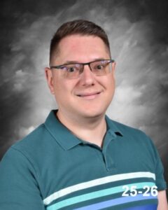 A smiling man with short hair and glasses, wearing a teal shirt with stripes, against a gray background.