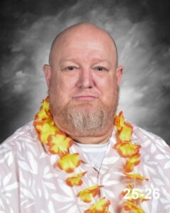 A man with a beard wearing a floral shirt and a yellow lei, set against a gray background.