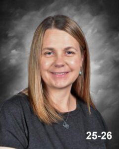 A smiling woman with long hair, wearing a dark top, against a gray background. She is looking directly at the camera.