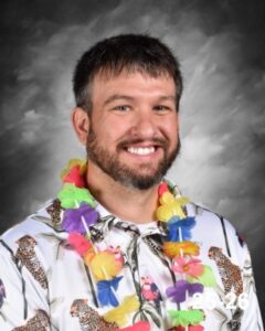 A smiling man with a beard wears a colorful Hawaiian shirt and a lei, posing against a gray background.