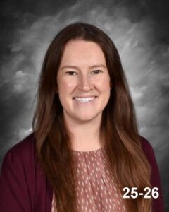 Smiling woman with long brown hair, wearing a maroon cardigan over a patterned blouse, against a gray background.