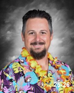 A man with a beard smiles, wearing a colorful Hawaiian shirt and a lei, against a grey background.
