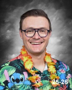 Smiling man wearing glasses and a colorful Hawaiian shirt with a floral lei against a dark background.