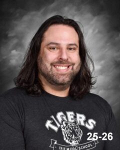 A smiling man with long hair wearing a black t-shirt featuring a tiger logo, against a gray background.