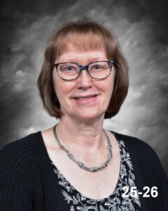 A woman with short brown hair and glasses smiles, wearing a black cardigan and a floral top, against a gray backdrop.