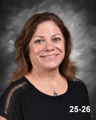 A smiling woman with long brown hair, wearing a black shirt, against a gray background. Text: "25-26" in the corner.