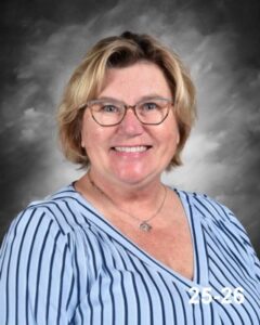 A smiling woman with short blonde hair wearing glasses and a striped blue blouse against a gray background.