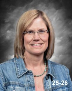 A middle-aged woman with shoulder-length blonde hair, wearing glasses and a denim jacket, smiles against a gray background.