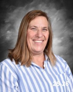 A woman with shoulder-length brown hair smiles, wearing a blue and white striped shirt, against a gray background.