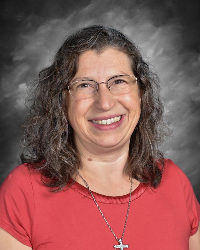 Mujer sonriente con gafas, que lleva una camiseta roja y un collar con una cruz, sobre un fondo gris.