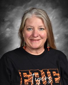 A woman with long hair, wearing a black shirt with "Tigers" text and decorative earrings, smiles against a gray backdrop.