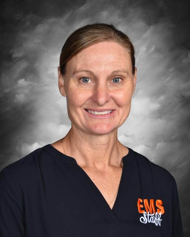 Mujer con una camiseta oscura del personal de emergencias médicas, sonriendo sobre un fondo gris.