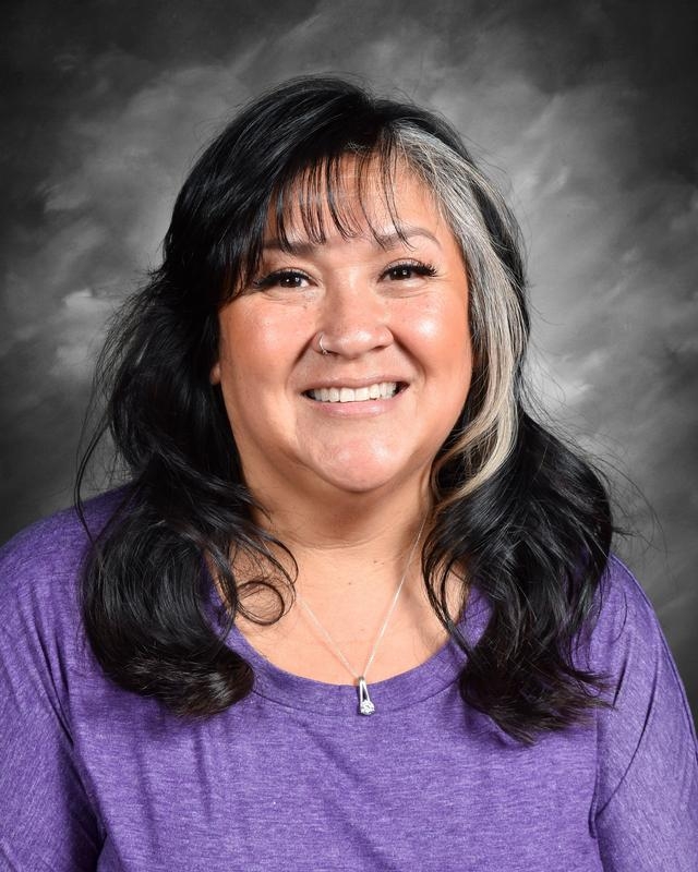 Mujer sonriente con el pelo largo y oscuro y una camiseta morada, sobre un fondo gris.