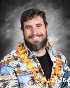 Smiling man with a beard wearing a colorful Hawaiian shirt and a flower lei, set against a gray background.