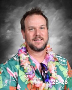 A man with a beard smiles, wearing a colorful Hawaiian shirt and a floral lei against a gray backdrop.
