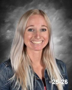 A smiling woman with long, light blonde hair wearing a denim jacket, set against a cloudy gray background.