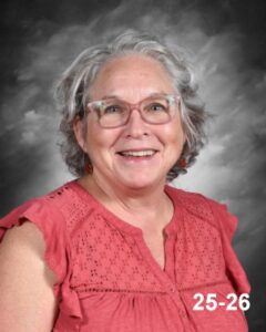 Smiling woman with gray hair and glasses, wearing a pink blouse, set against a gray background.