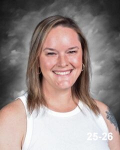 A smiling woman with long brown hair and a tattoo, wearing a white tank top, against a gray background.