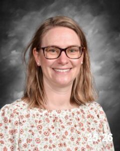 Smiling woman with shoulder-length hair wearing glasses and a floral blouse, set against a gray background.