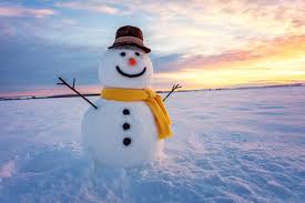  A cheerful snowman stands in a snowy field during sunset. The snowman has a brown hat, a bright yellow scarf, and stick arms spread wide. Its face has a smiling carrot nose, two black eyes, and a row of coal buttons. The background shows a colorful sky blending orange, pink, and blue hues over a vast, untouched blanket of snow.