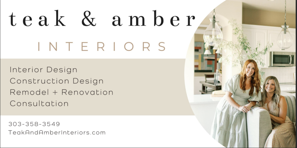 A business card for "Teak & Amber Interiors," an interior design company. The card features a clean, modern design with a neutral color palette of beige, white, and soft brown. The company's name is written in a stylish serif font, with "INTERIORS" in a lighter sans-serif font below. Listed services include Interior Design, Construction Design, Remodel + Renovation, and Consultation. The contact details, including a phone number (303-358-3549) and website (TeakAndAmberInteriors.com), are displayed at the bottom. On the right side of the card, there is a photograph of two women smiling in a beautifully designed kitchen. One woman has long red hair and wears a soft green dress, while the other has long blonde hair and wears a patterned sleeveless dress. They are leaning on a light gray sofa with a relaxed and welcoming posture. The background features a modern, well-lit kitchen with white cabinets, glass pendant lights, and greenery