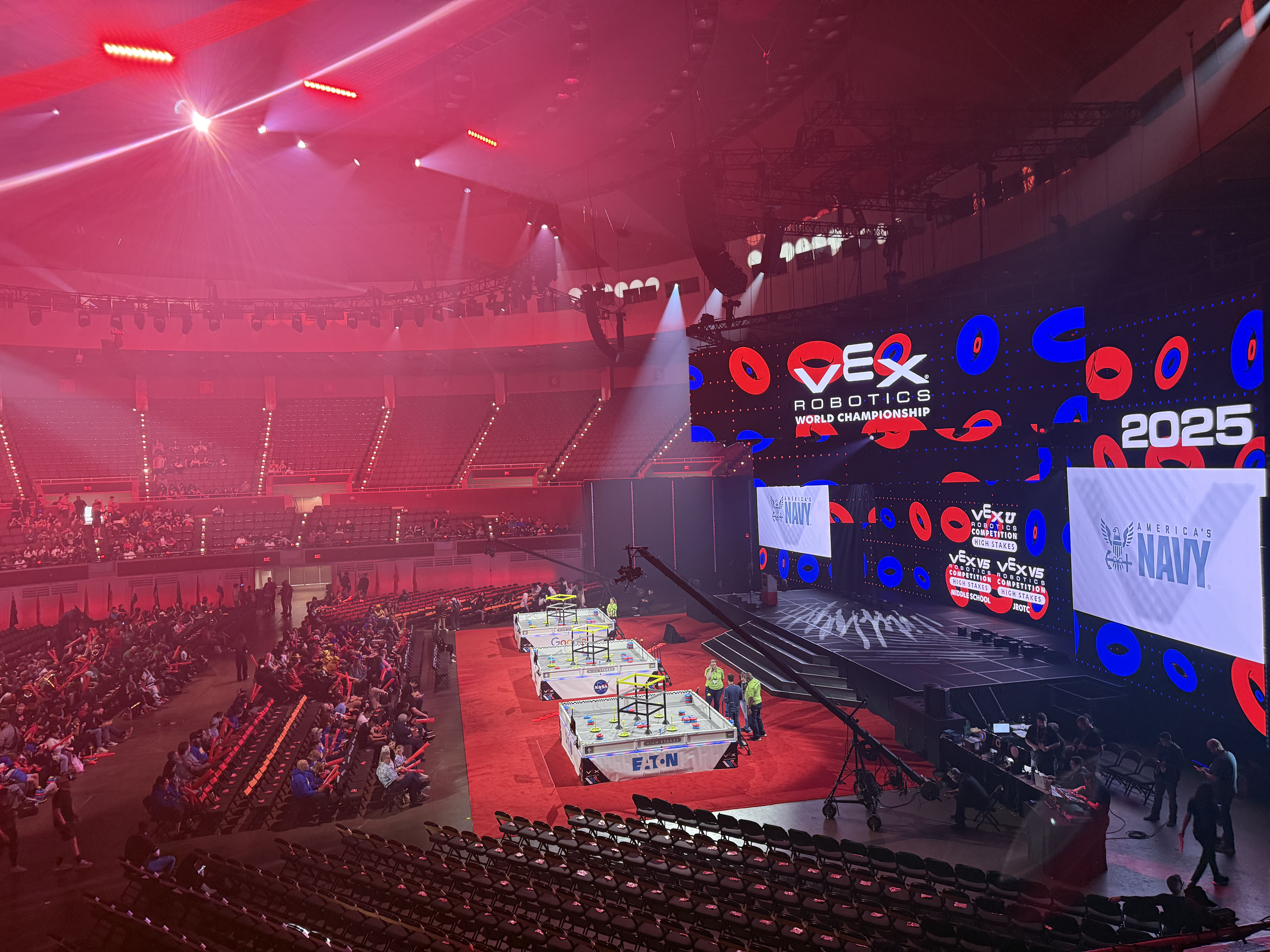 The image shows a large indoor arena hosting the 2025 VEX Robotics World Championship. The stage at the far end features massive LED screens displaying the VEX Robotics logo, the year 2025, and a sponsorship banner for America’s Navy. In the foreground, there are multiple robotics competition fields set up, surrounded by participants and event staff. The arena is partially filled with spectators, and red lighting adds a dramatic effect to the venue. A crane camera is positioned near the competition fields for live event coverage. The overall atmosphere is energetic and high-tech, fitting for a world championship robotics event.