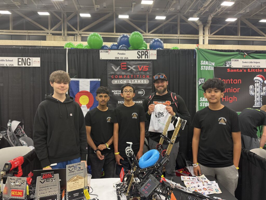 Five individuals stand at a robotics event booth, showcasing their team's achievements and holding a T-shirt.