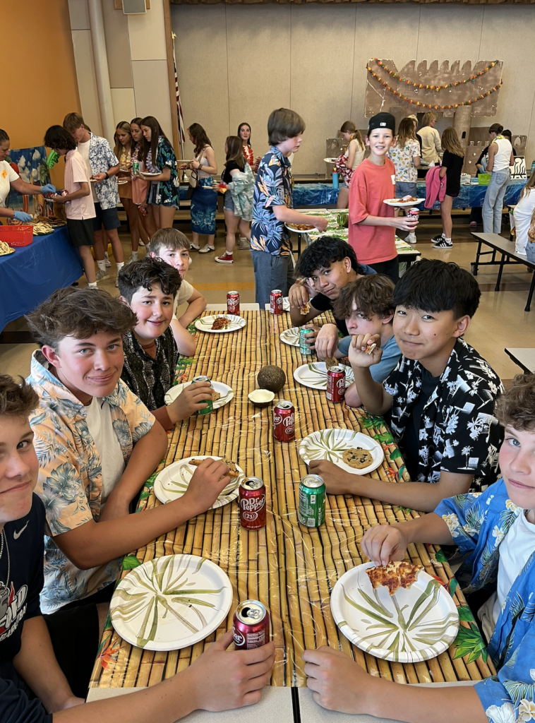 A group of boys sits at a table decorated with tropical print, enjoying food and drinks at a festive gathering.