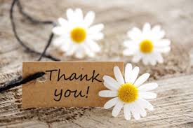 A close-up of a rustic wooden surface with three white daisies and a small brown tag tied with black string. The tag reads 'Thank you!' in cursive writing.