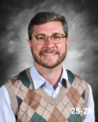 A smiling man with glasses and a patterned sweater vest poses against a gray backdrop. He appears friendly and approachable.
