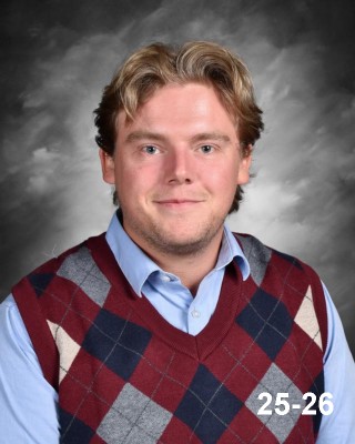 A young man with wavy hair smiles, wearing a blue shirt and a maroon and navy argyle sweater vest.