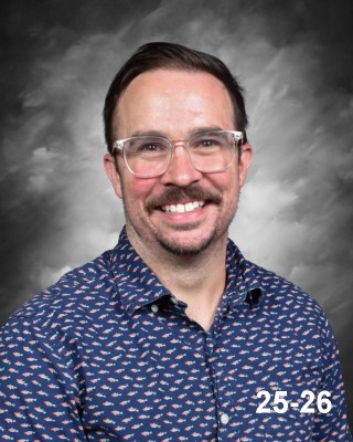 A smiling man with glasses and a patterned blue shirt poses in front of a gray backdrop. He has a mustache.