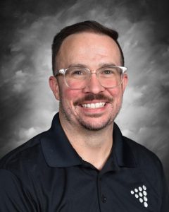 Smiling man with glasses and a beard, wearing a black shirt, against a gray studio background.