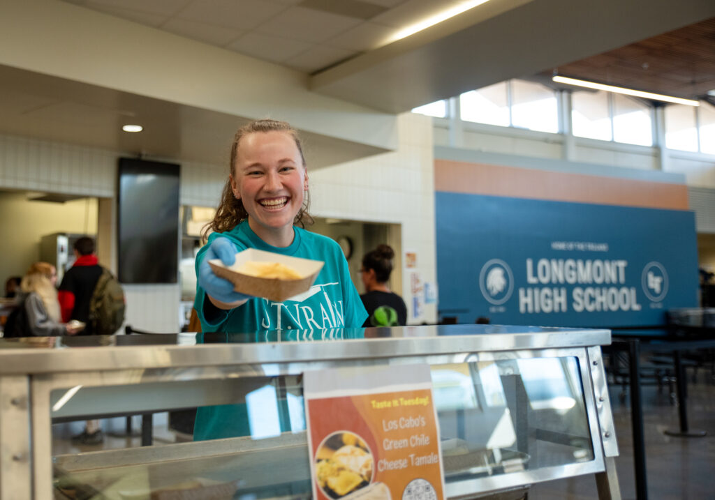 A smiling student in a blue shirt hands out food at Longmont High School's cafeteria.