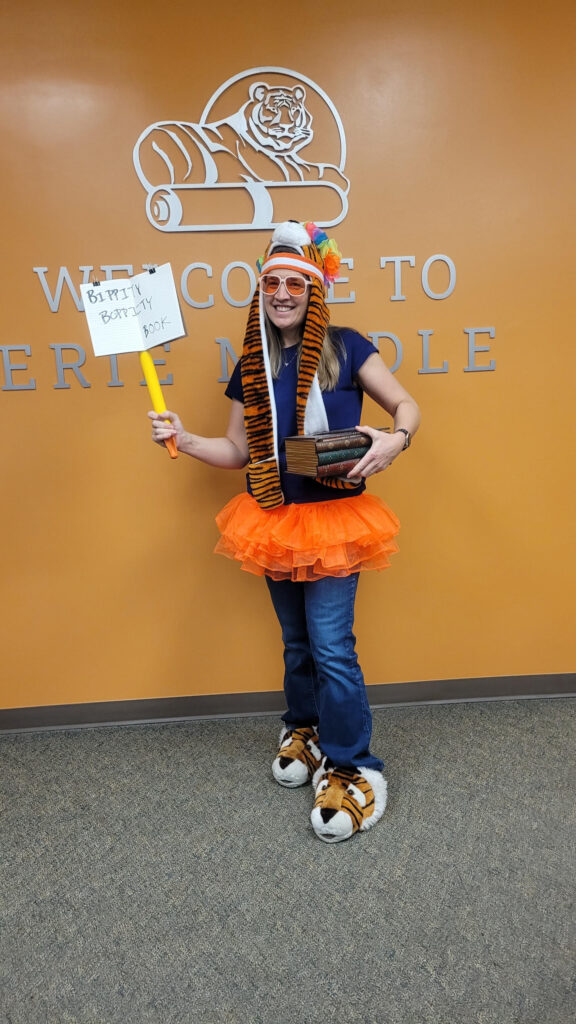 A woman in a colorful outfit holds a sign and books in front of a school welcome sign, smiling and dressed playfully.