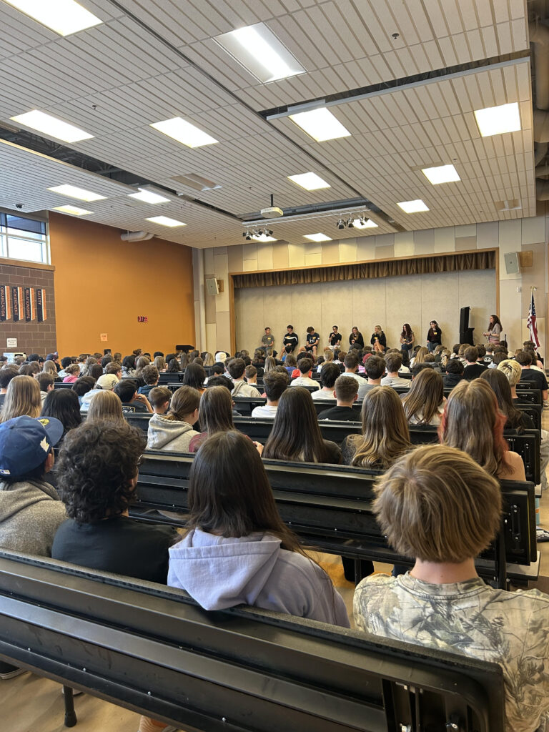 A crowded auditorium with students seated, facing a stage where several speakers are presenting.