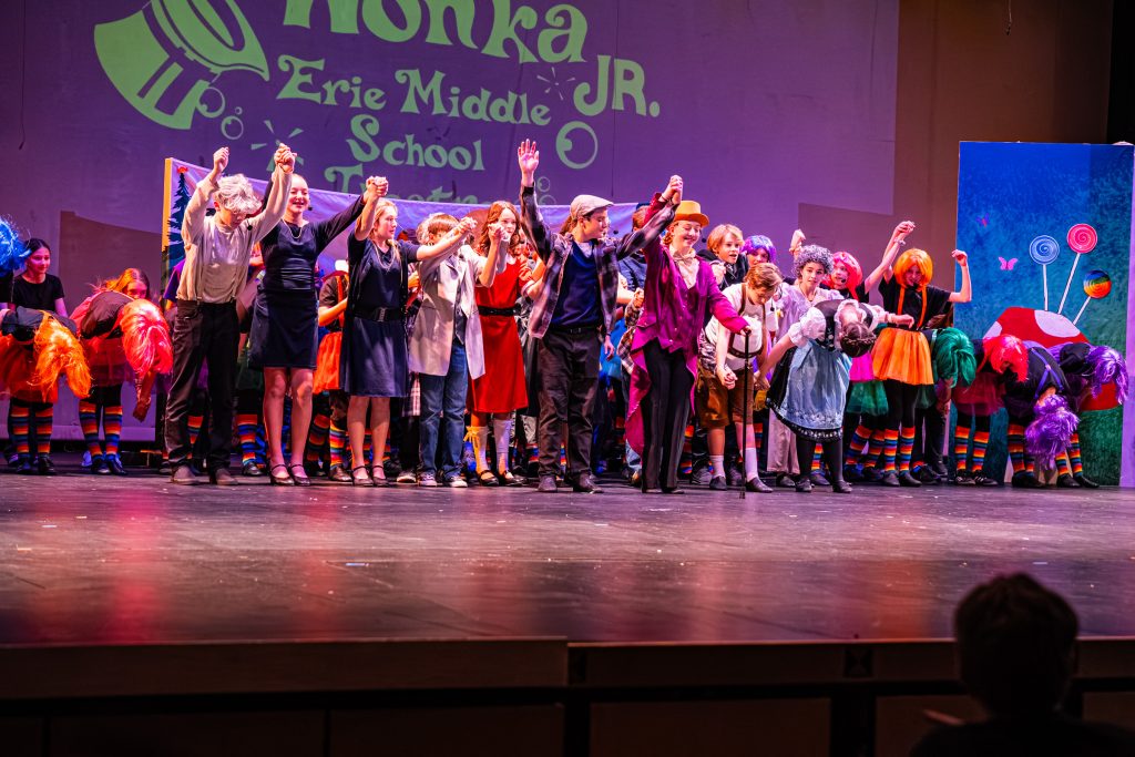 A group of students on stage at Erie Middle School bows after a performance of "Willy Wonka Jr." in colorful costumes.