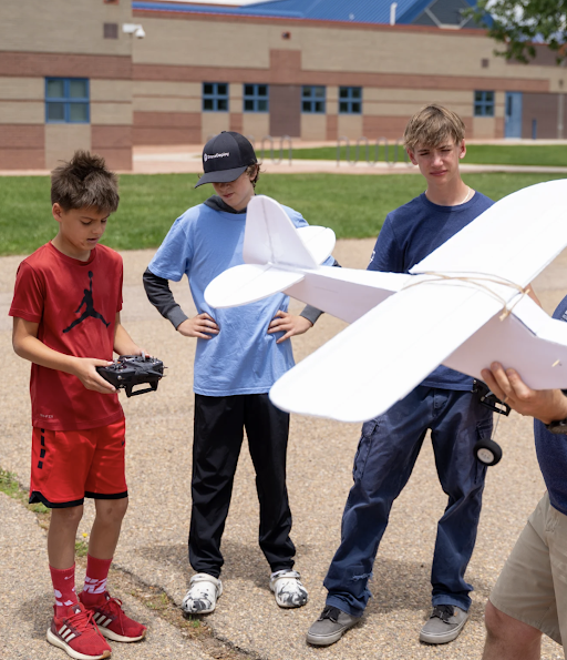 Three boys observe as one holds a remote control while another prepares a model airplane for flight outdoors.