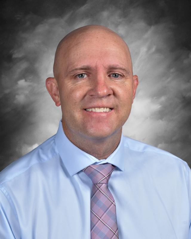 Un hombre con camisa azul claro y corbata a cuadros rosas, sonriendo, con un fondo gris nublado.